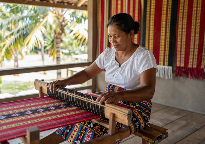 East Timorese woman weaving traditional tais cloth on a wooden loom with vibrant red gold and black textile patterns